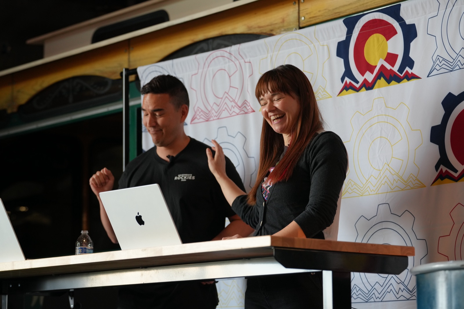 Two organizers laughing together on stage in front of a Colorado flag backdrop