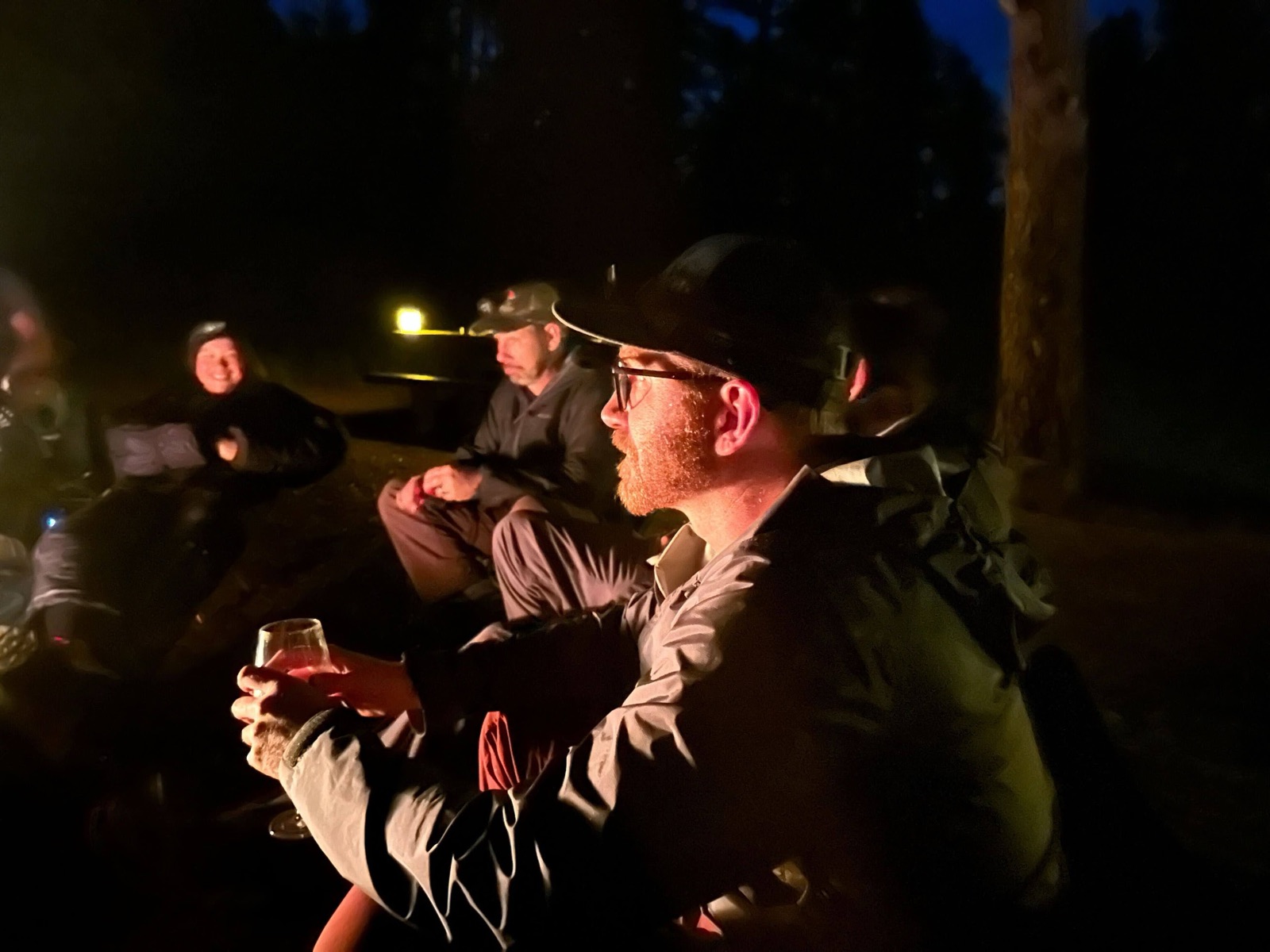 A group gathered around a campfire at night, sharing drinks and conversation